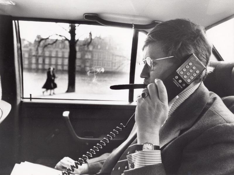 Minister Hans Wiegel met een autotelefoon in de dienstauto; op de achtergrond het Binnenhof